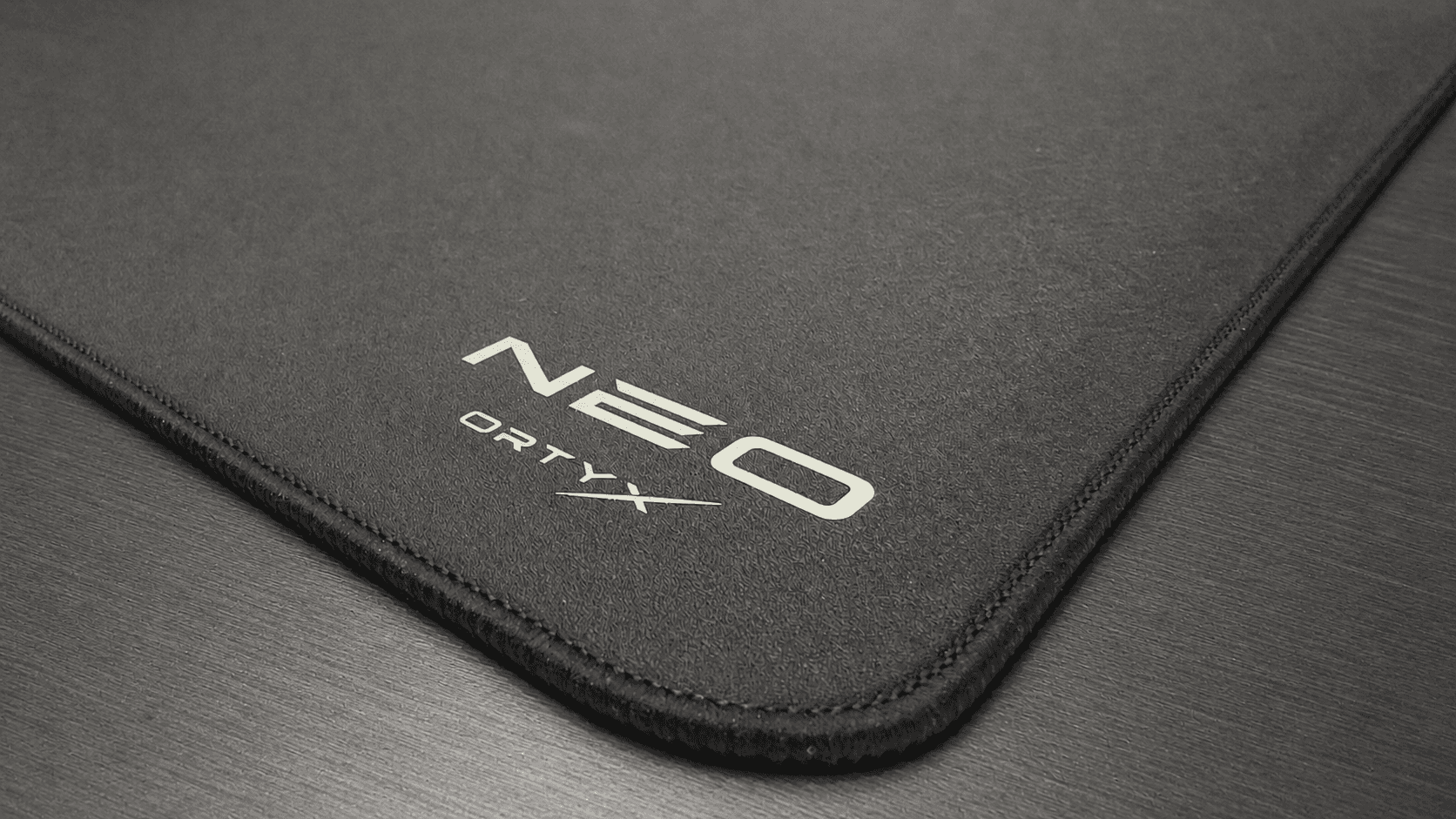 The corner of the ORTYX NEO mousepad on a dark desk, its stitched edge and printed NEO ORTYX mark catching a low raking light